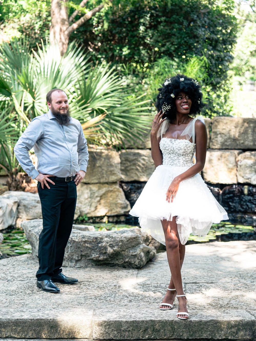A couple posing on garden steps, with a white short dress and light blue shirt against lush foliage.