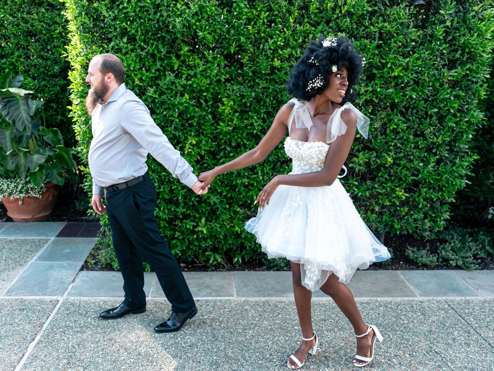 A couple dance playfully in front of a green hedge, with the bride wearing a short white dress and strappy heels.