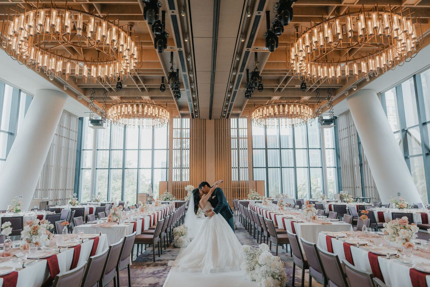 Two people embrace in an elegant wedding venue with crystal chandeliers and floor-to-ceiling windows overlooking city views.