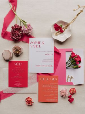 Wedding invitation suite in pink and coral tones with roses and gold jewelry on a beige background.