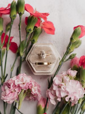 An engagement ring sits in a velvet ring box surrounded by pink carnations and red flowers with green stems.