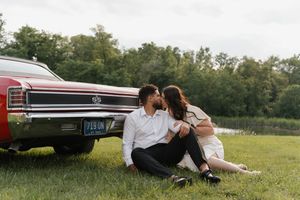 A couple sits together on grass beside a vintage red Chevelle car on a sunny summer day.