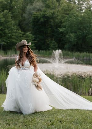 Western-inspired bride in flowing white gown and cowboy hat poses by pond with fountain in rustic outdoor setting.