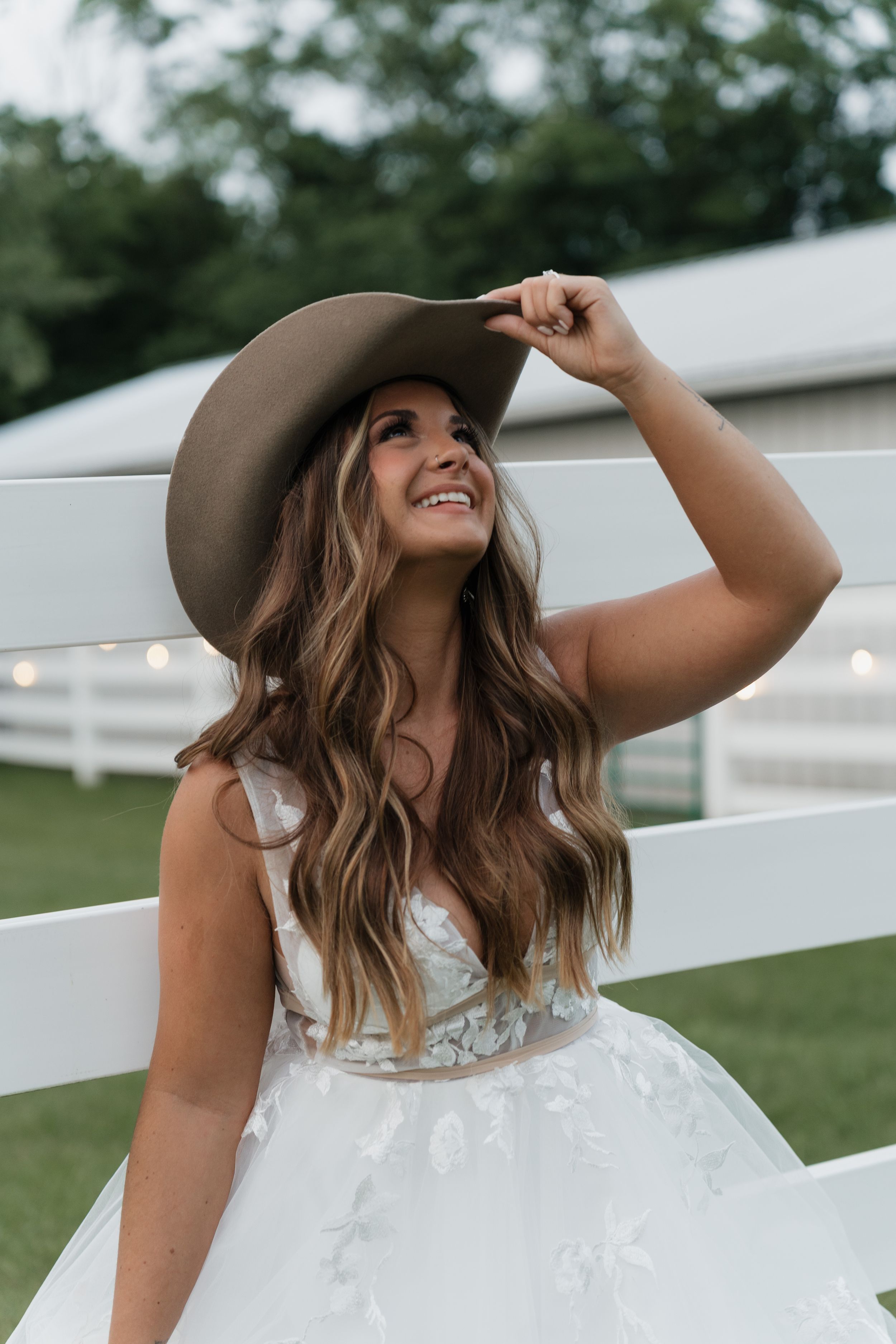 A bride in a white wedding dress and brown cowboy hat stands by white fence on her wedding day with a joyful smile.