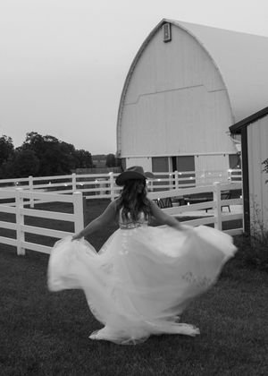 A twirling figure in a white wedding dress stands in front of a classic white barn and white fence on a country farm.