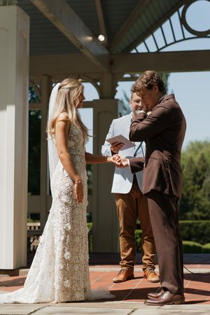 A wedding ceremony takes place outdoors at a gazebo with the bride wearing a detailed lace gown and officiant standing nearby.