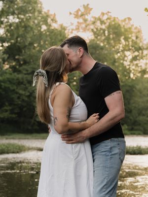 A romantic couple embraces in a white dress and casual attire during sunset in a serene outdoor setting by a lake.