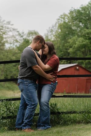 A couple shares a romantic moment by a rustic red barn and wooden fence in a rural countryside setting.