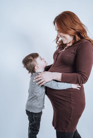 Tender moment between expectant mother in brown dress and young child against white background.