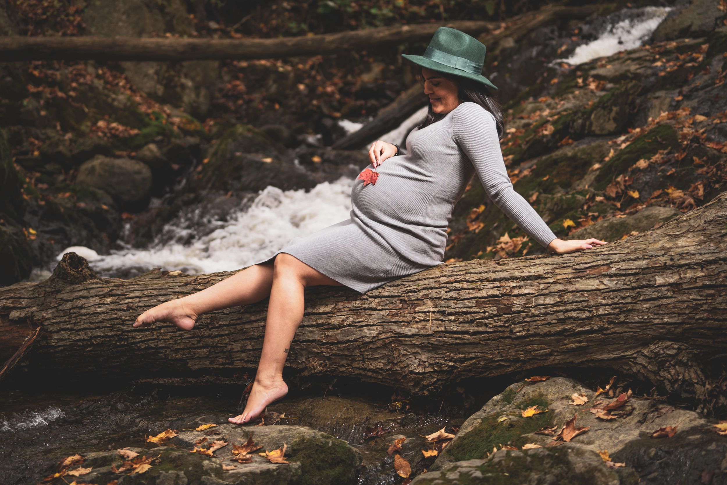 Maternity photo of a woman in gray dress relaxing on a fallen log near a stream.