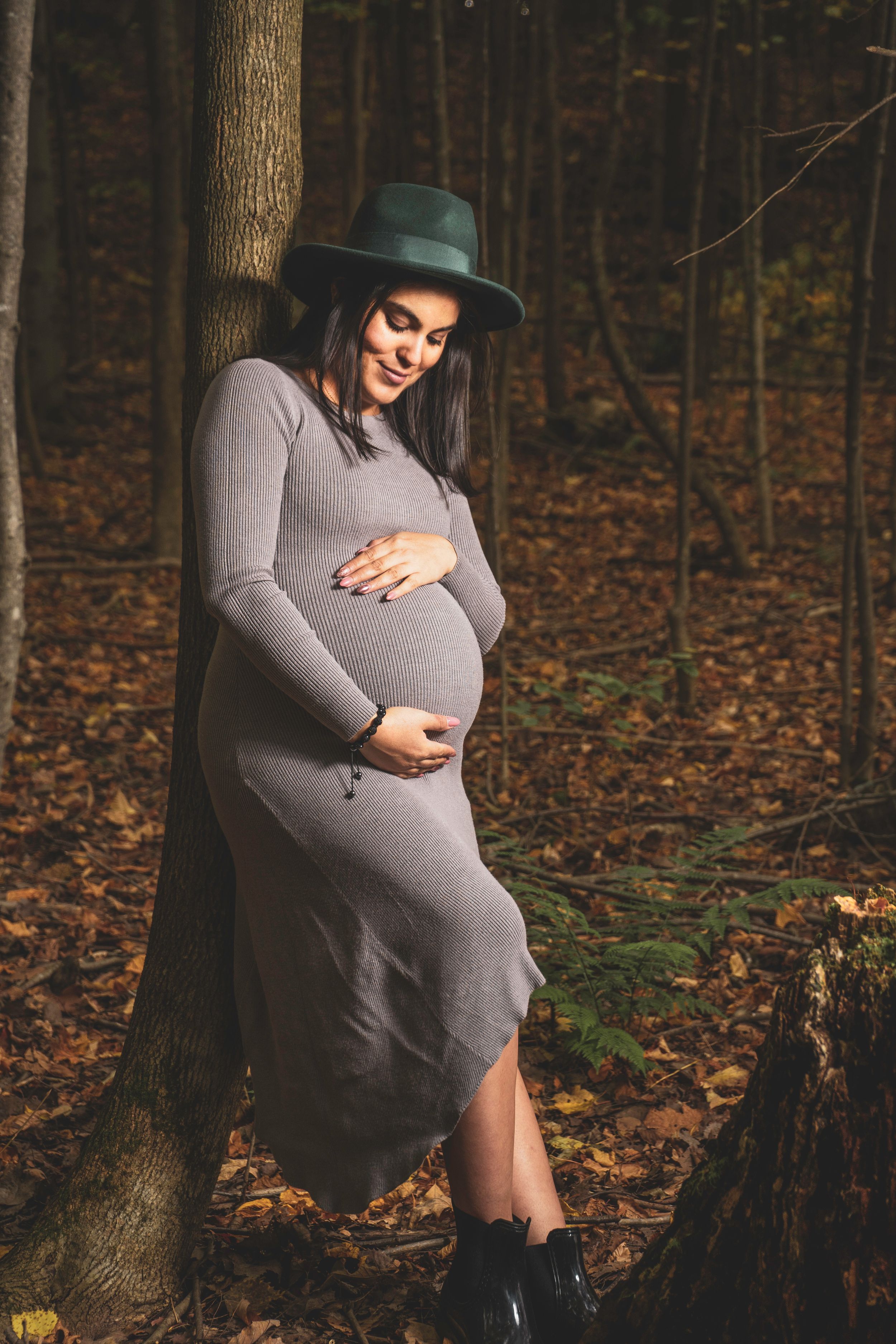 Expectant mother in a gray dress and green hat poses thoughtfully in an autumn forest setting.