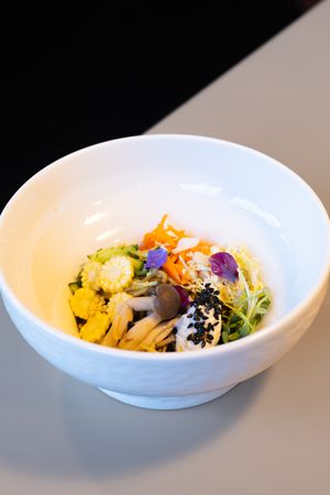 Artfully plated rice bowl with colorful vegetables, edible flowers, and black sesame seeds in a white ceramic dish.
