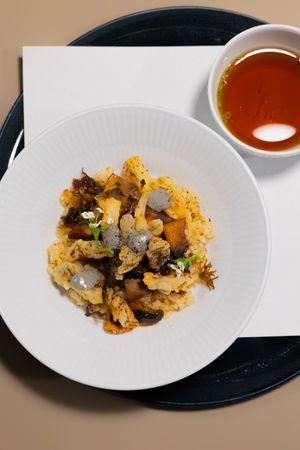 A roasted cauliflower dish with mushrooms and herbs served with a side of dipping sauce on a white plate.