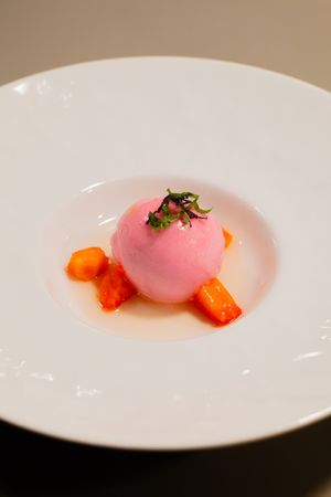 A pink spherical dessert with orange garnish and microgreens on a white plate.