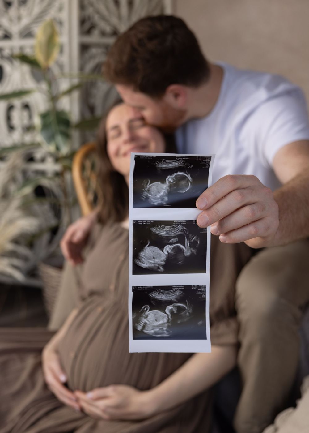 A series of black and white ultrasound photos being held up against a cozy home setting with plants in the background.