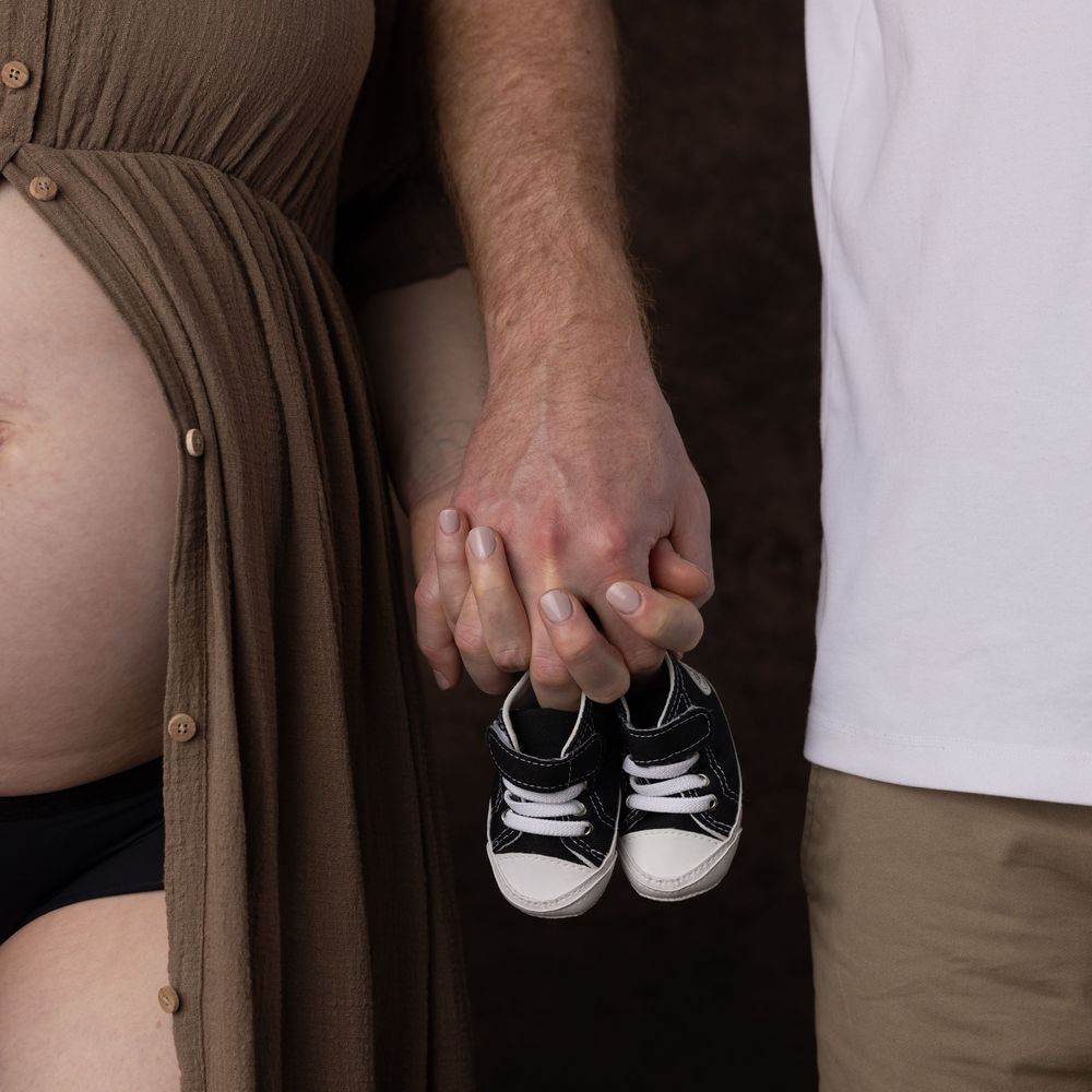 A close-up of hands holding tiny black and white baby shoes next to a pregnant belly in a brown dress.