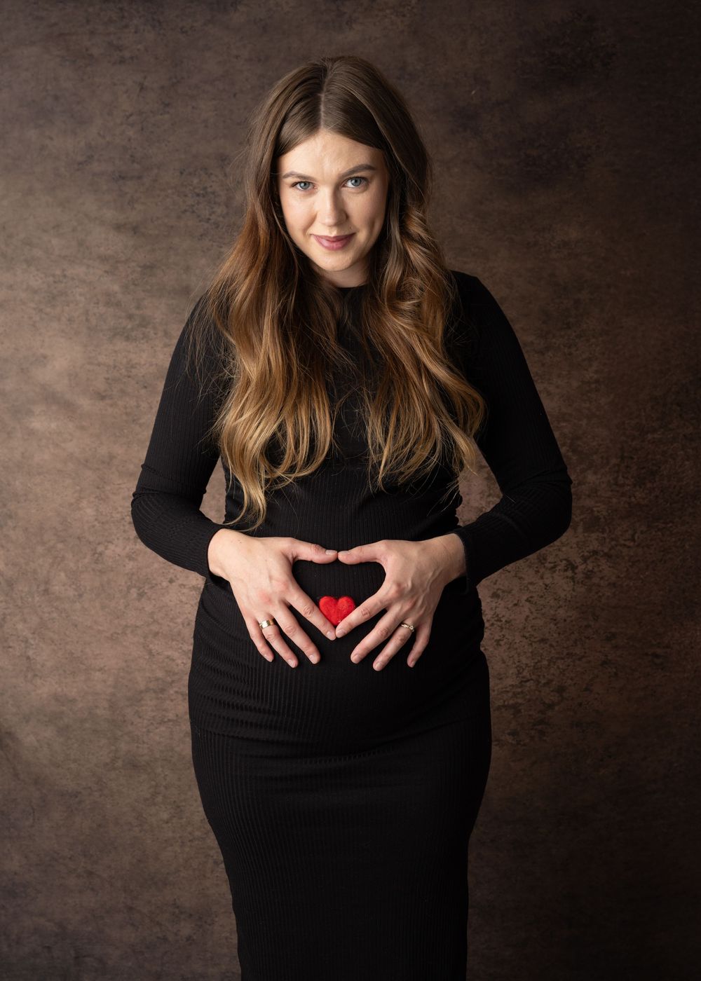Studio portrait of someone in a black dress making a heart shape with hands over their stomach against a brown backdrop.