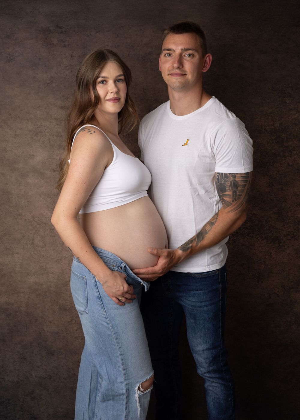 A couple in white tops and jeans poses for a maternity photo shoot against a dark textured backdrop.