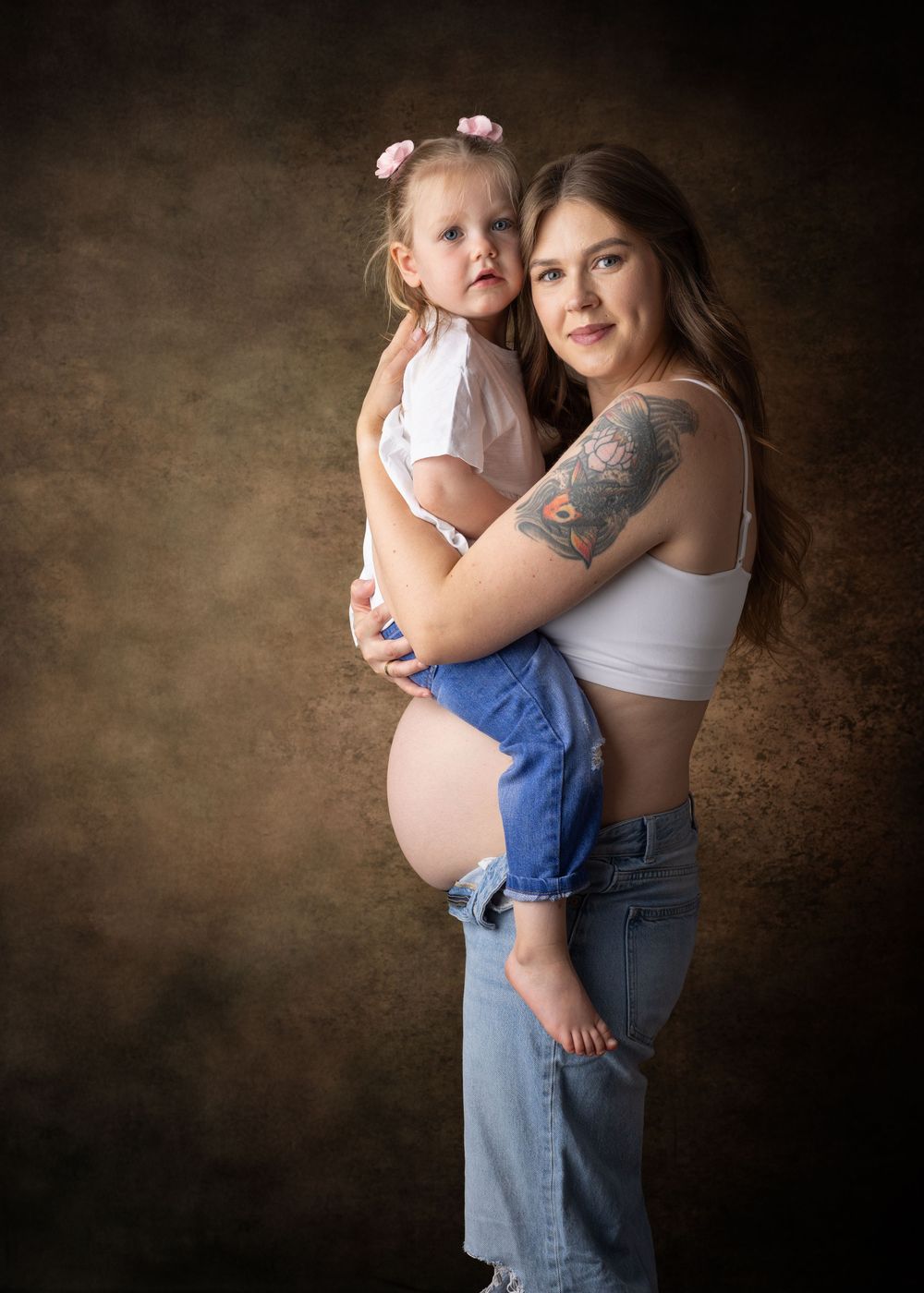 A series of intimate maternity portraits showing a woman in jeans and white top holding a small child against a dark backdrop.