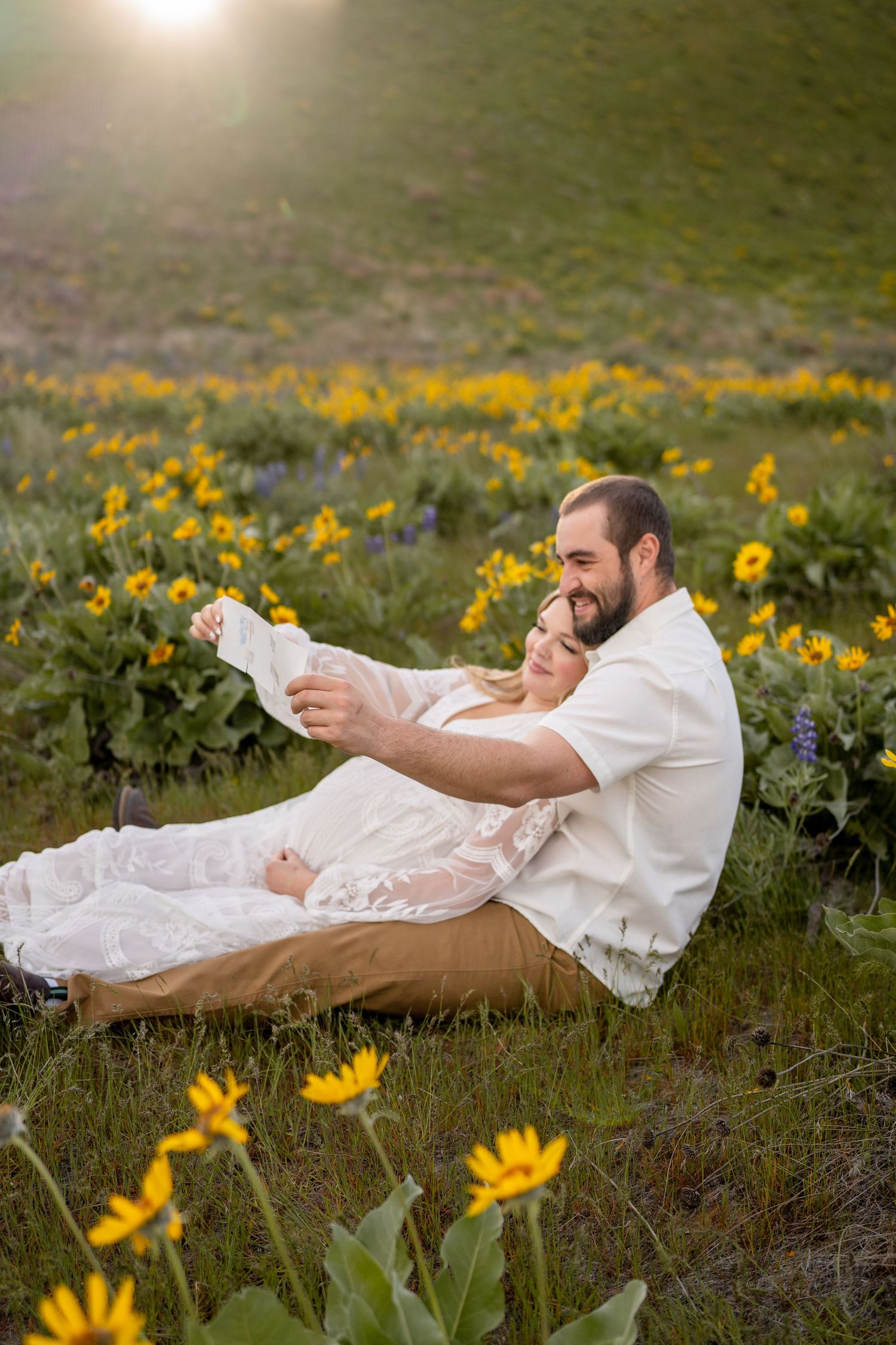 A couple sits together in a field of yellow wildflowers during sunset, reading a book together.