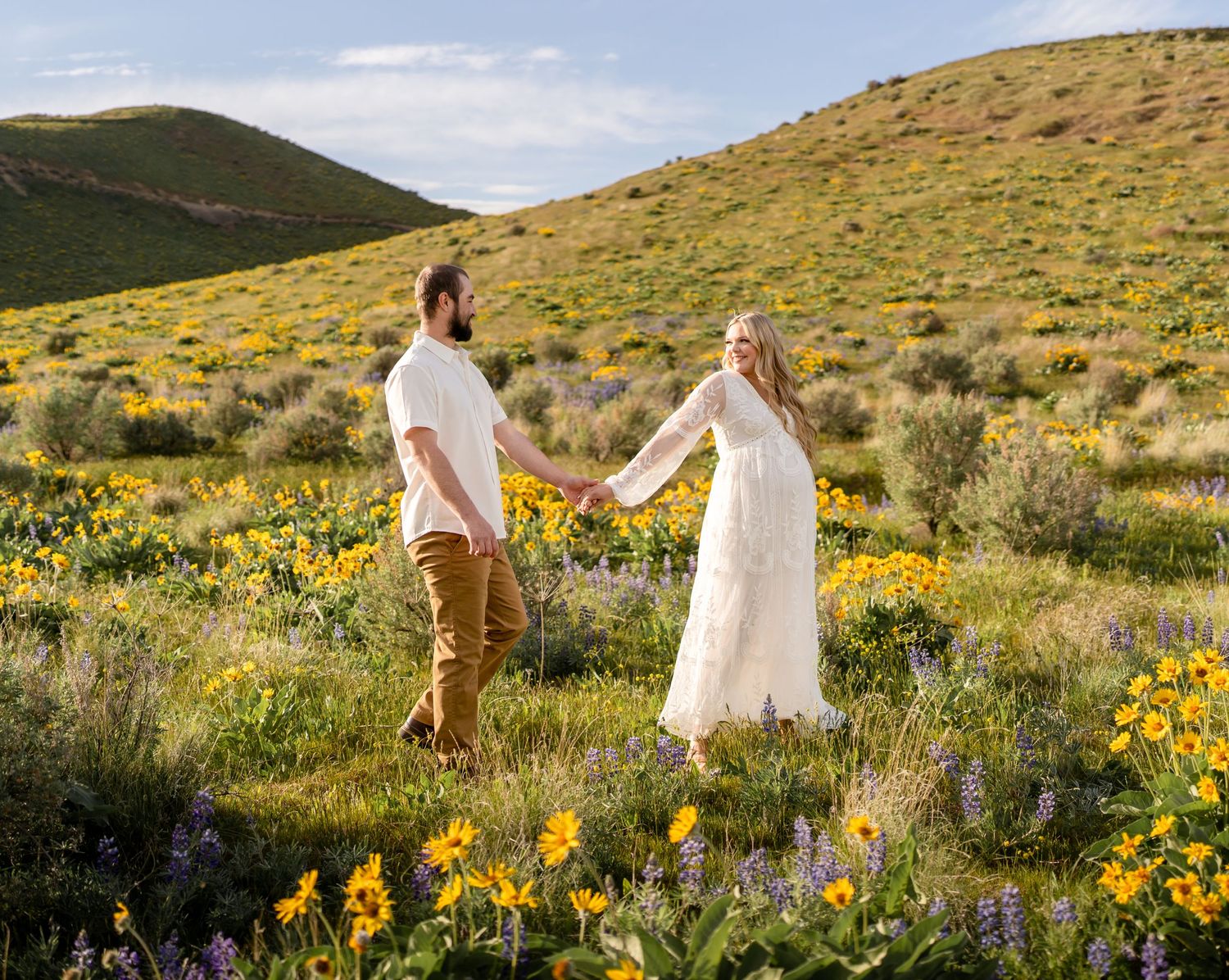 Couple holding hands while walking through a mountainside meadow filled with yellow wildflowers.