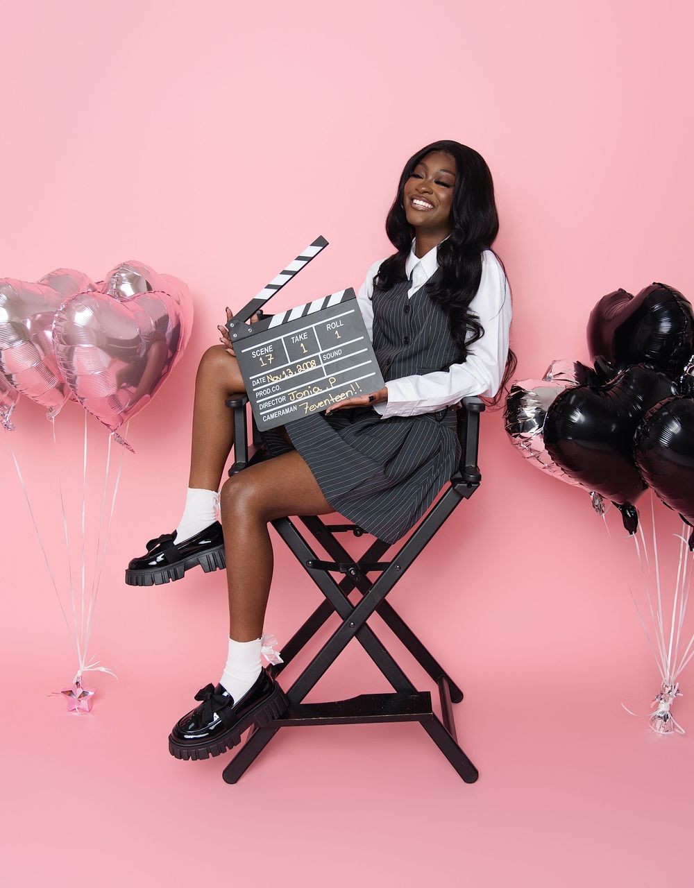 Stylish student sitting in director's chair with movie clapboard against pink background with silver and black balloons.