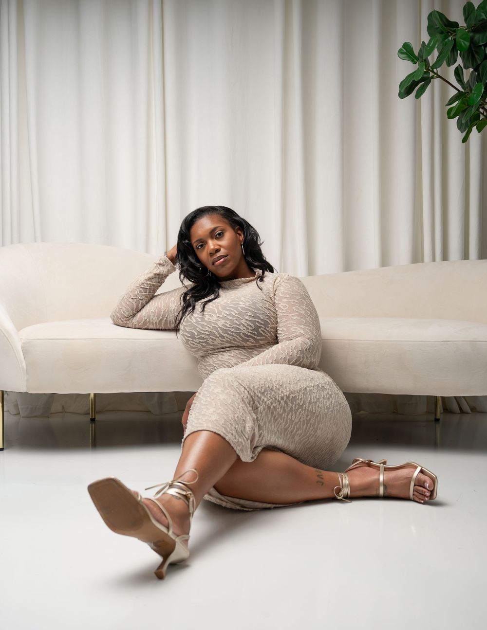 Woman in beige long-sleeve mini dress and gold strappy heels poses on white floor beside cream sofa.