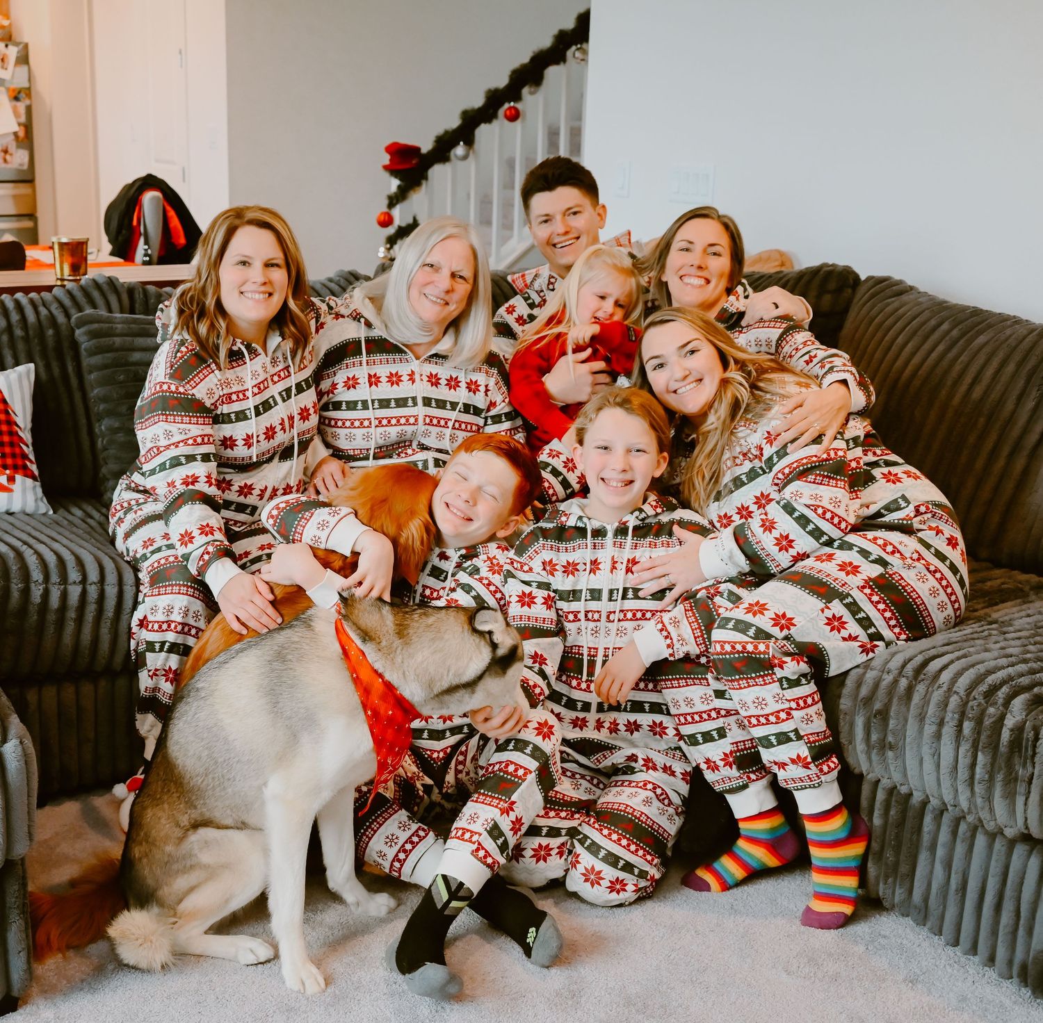 A group in matching holiday pajamas sits together on a grey couch with their white dog during Christmas festivities.