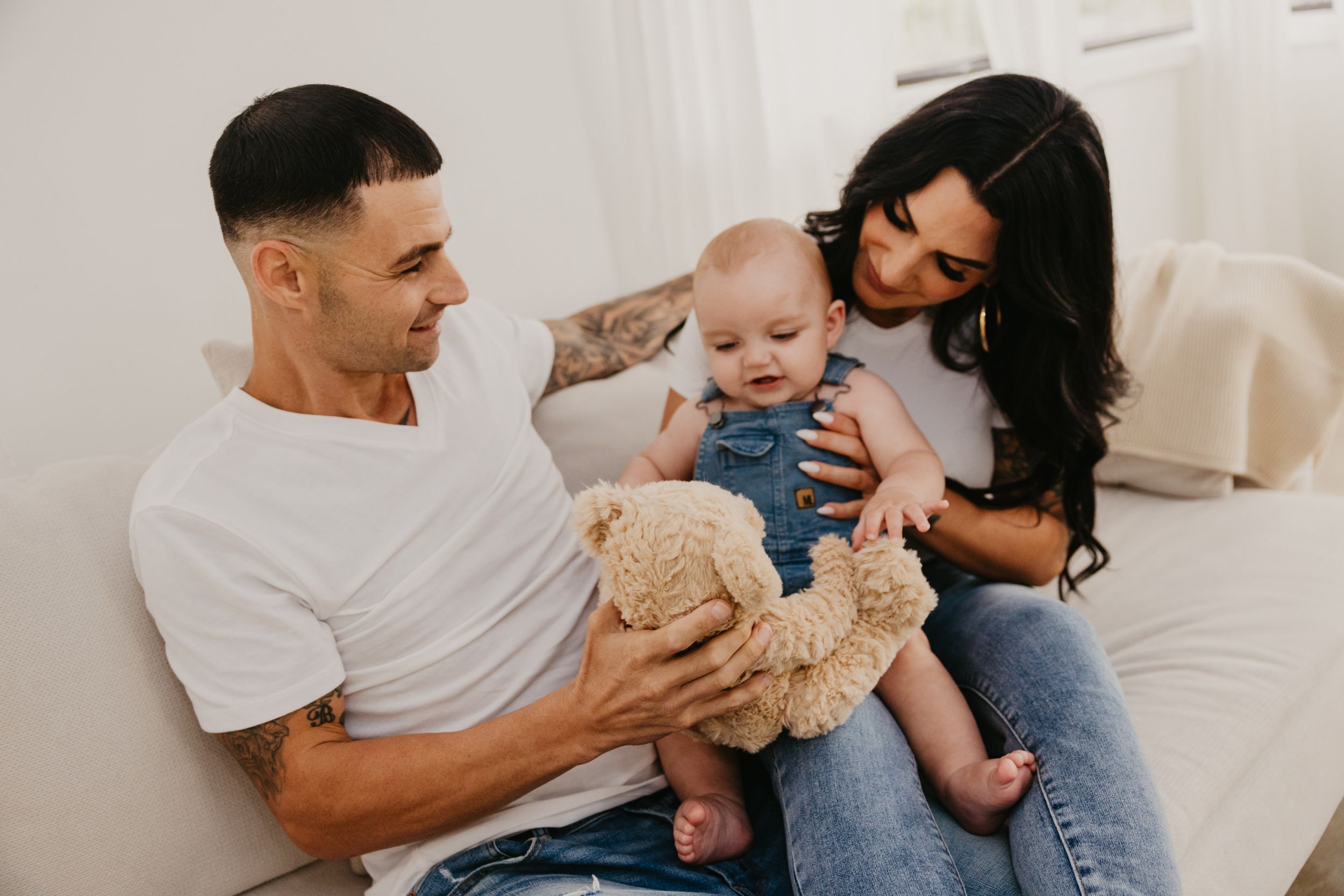 Family shares playful moment with baby and teddy bear on white couch.