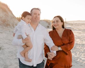 A family wearing white and rust-colored clothing poses together on a sandy beach at sunset.
