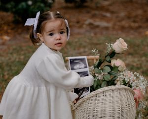 A small child in a white dress stands near a wicker basket filled with flowers and a framed photo on autumn ground.