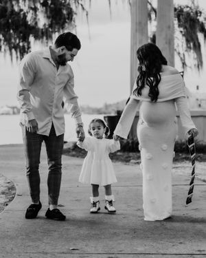 A black and white photo of a loving family moment showing parents standing with a small child in a white dress between them.