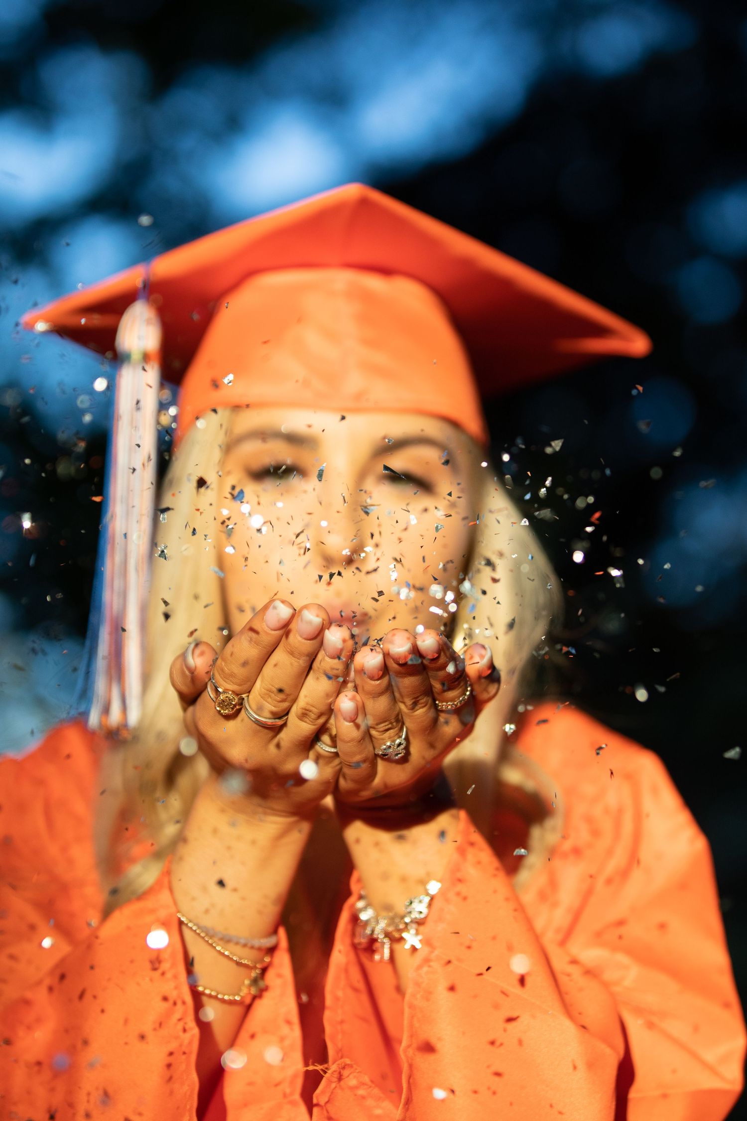 Orange graduation cap with glitter being tossed in celebration against blue bokeh background.