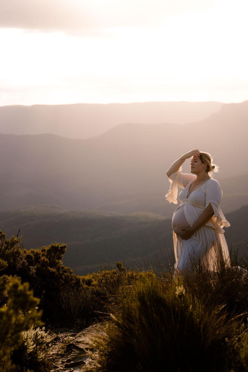 Pregnant woman in flowing white dress moving gracefully against mountain landscape at dusk.