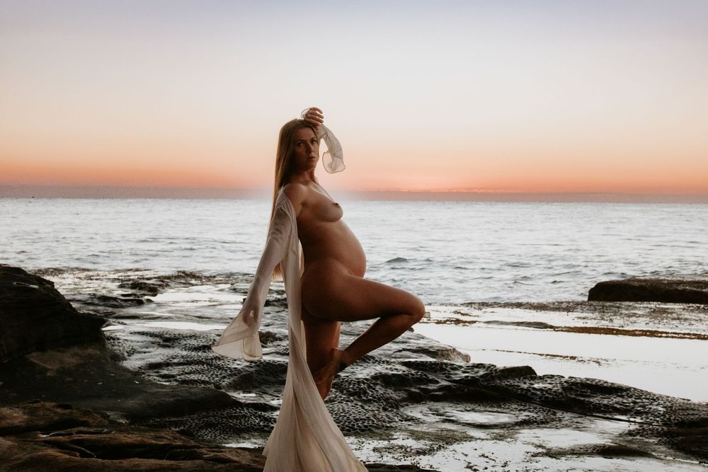 A silhouetted gorgeous pregnant maternity stands on rocky coastal cliffs at sunset while waves crash against the shore in the background.