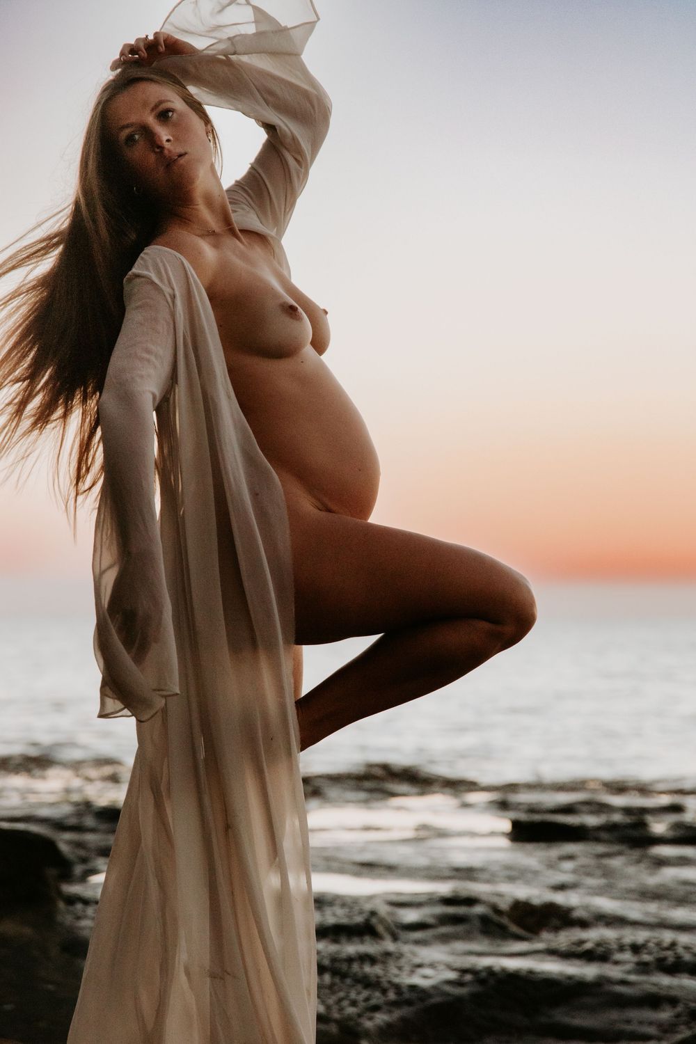 Artistic silhouette of a pregnant mama in flowing white fabric posing gracefully against a sunset ocean backdrop.