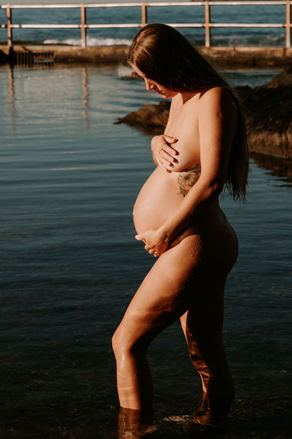 Silhouetted figure stands in shallow ocean water during sunset, creating a serene maternity photoshoot scene.