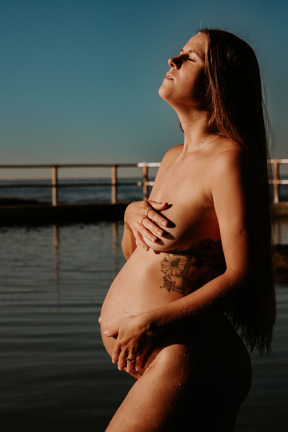Nude pregnant woman with eyes closed, hands on her bump, sunlight warming her face, captured in serene natural light at North Curl Curl Beac