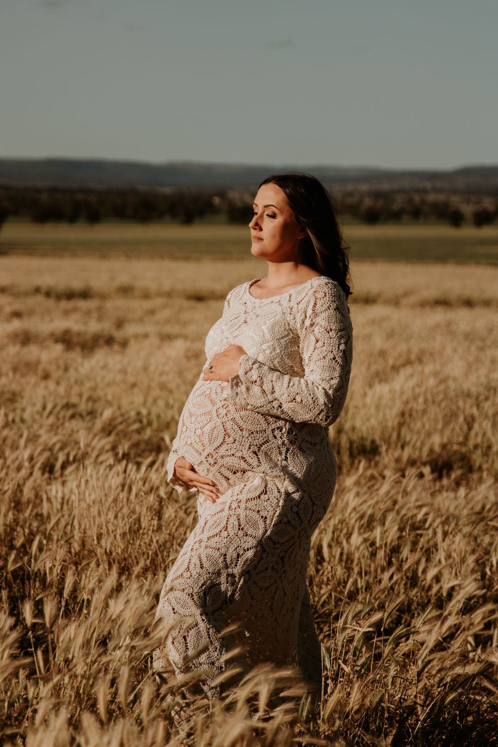Maternity photoshoot of woman in lace dress standing in golden wheat field at sunset.