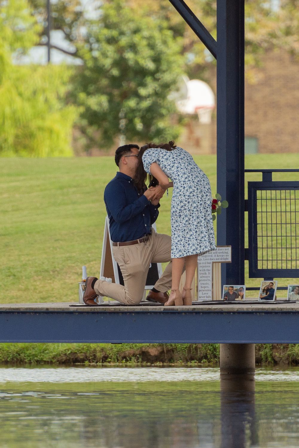 A man kneels on a dock while proposing, holding his new fiancée's hands as she bends over in disbelief, full of emotions.
