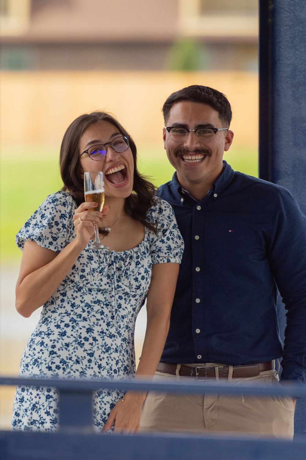 A woman smiles big while holding a champagne glass in excitement next to her new fiancé who laughs joyfully.