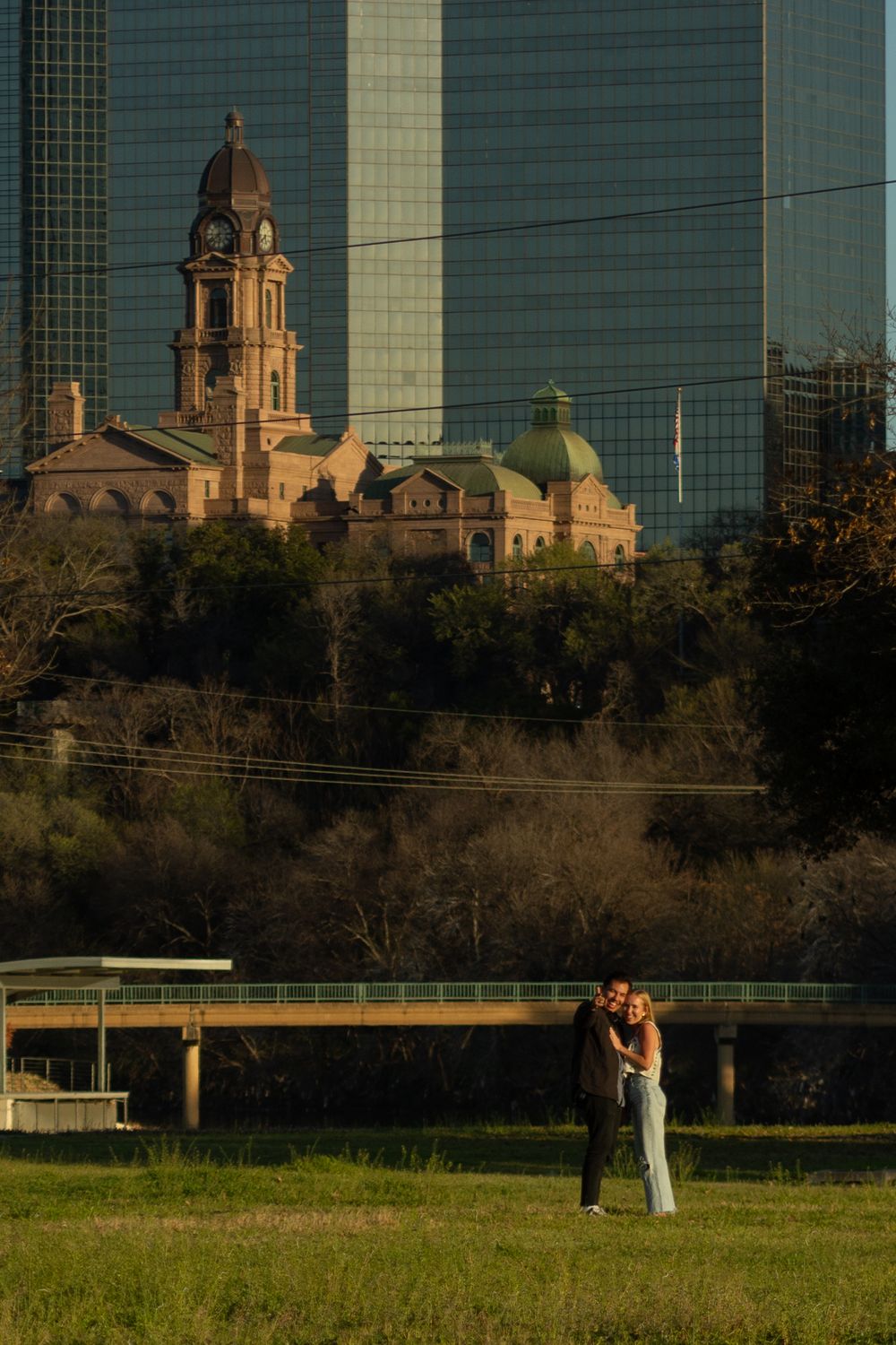 Faraway photo of a couple standing and smiling at the camera at sunset in a Fort Worth park with the downtown buildings in the background.