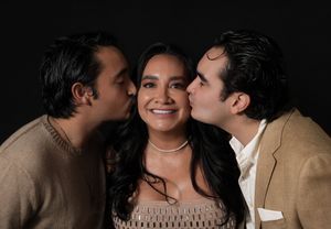A smiling woman receives kisses on her cheeks from two men in a warm, affectionate portrait against a dark background.
