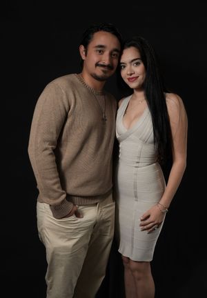 A young couple poses together against a dark background, man in beige sweater and woman in white dress.