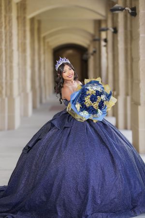 Young woman in navy blue quinceañera ball gown with crown holds matching floral bouquet in elegant archway.