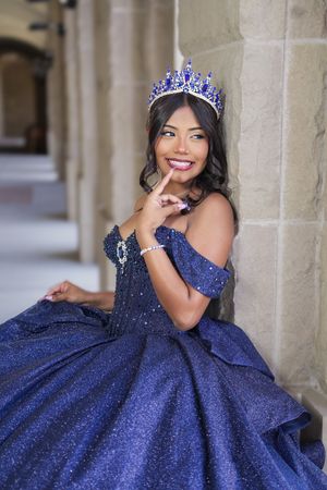 Young woman in sparkling blue quinceañera gown and jeweled crown smiles while posing against stone columns.