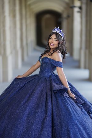 Young woman in a sparkling navy blue quinceañera ball gown and tiara poses gracefully in an elegant archway.