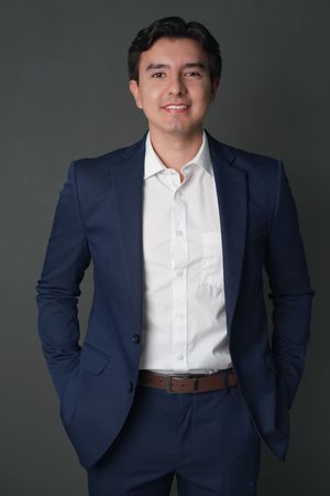 Young professional man in navy blue suit and white shirt smiling confidently against dark gray background.