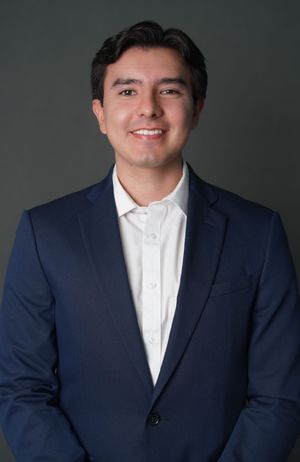 Young professional man smiling in navy blue blazer and white shirt against dark gray background.