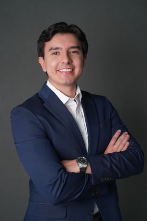 Professional man in navy suit with arms crossed, smiling confidently against a dark gray background.
